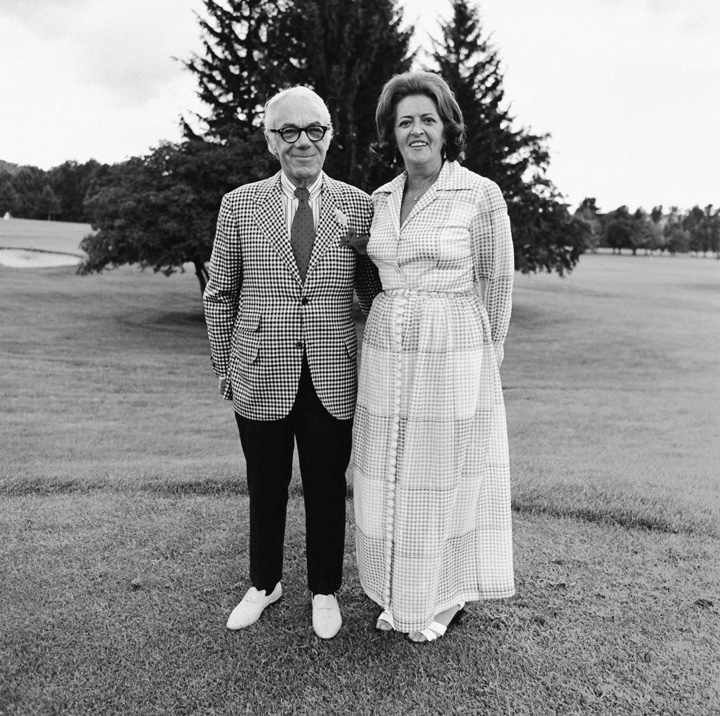 A black and white wide shot of a mature couple in formal attire standing on a golf course.