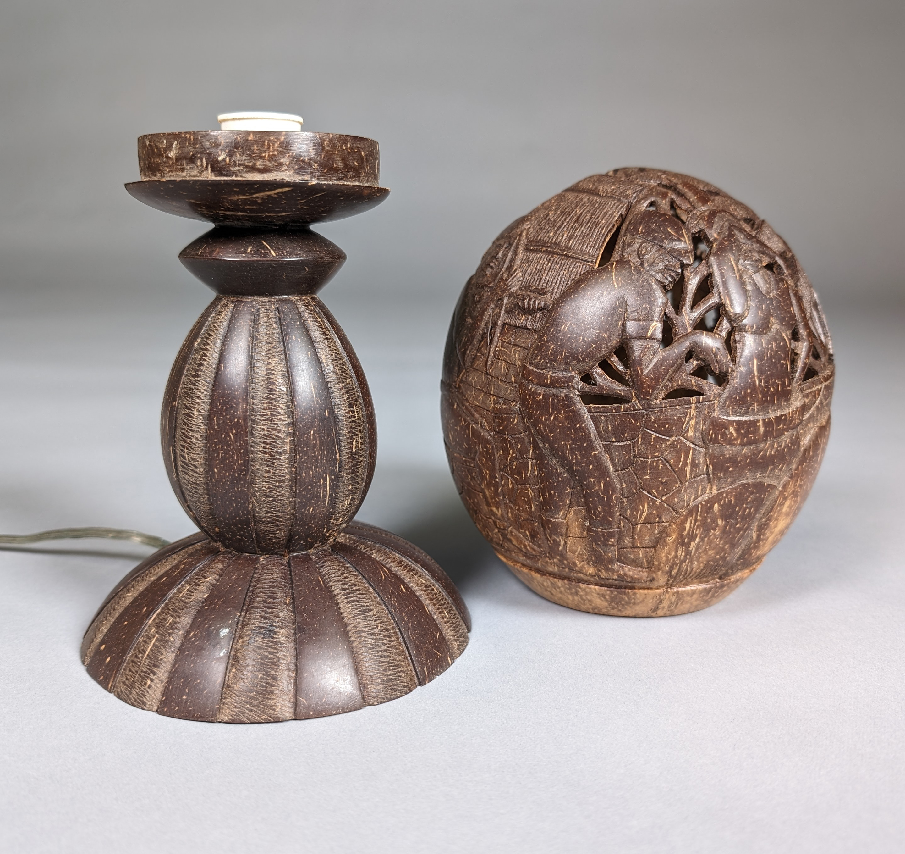 A crafted lamp made from a coconut shell, displayed alongside a carved coconut shell artwork. The lamp features a textured, grooved design, with the top portion holding a light fixture. The adjacent carved coconut shell depicts an intricate scene of people engaged in daily activities, highlighting detailed craftsmanship. Both items are showcased against a plain gray background.