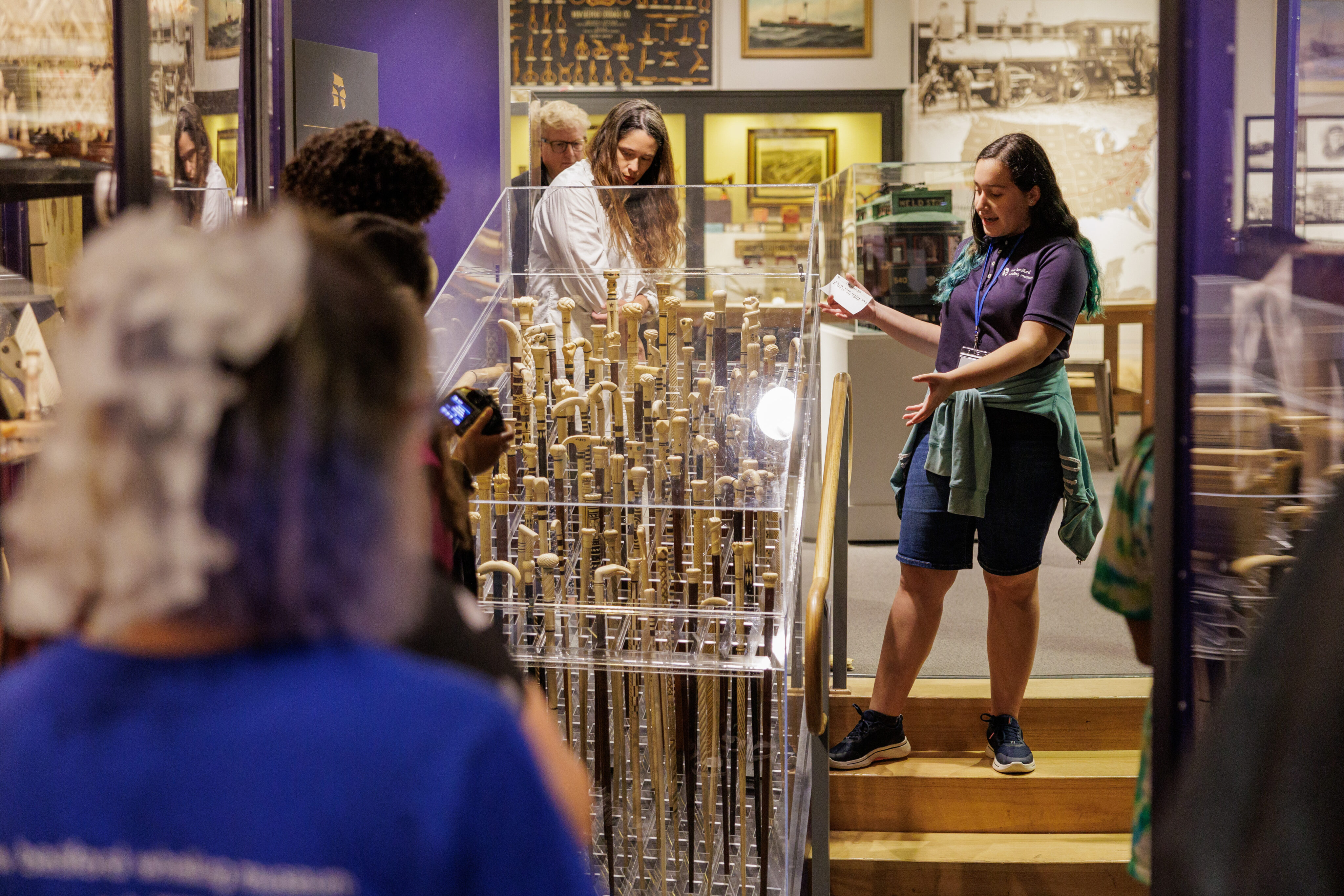 An apprentice gives a guided tour in the museum, focusing on a display of scrimshaw canes housed in a glass case. The apprentice explains the intricate craftsmanship to visitors.