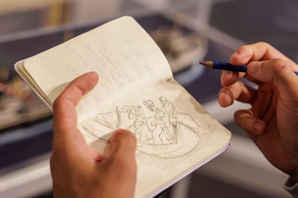 Close-up of a person's hands holding a sketchbook and pencil, featuring a detailed drawing of figures in a whaleboat. The sketch captures movement and detail, with faint notes written on the opposite page.