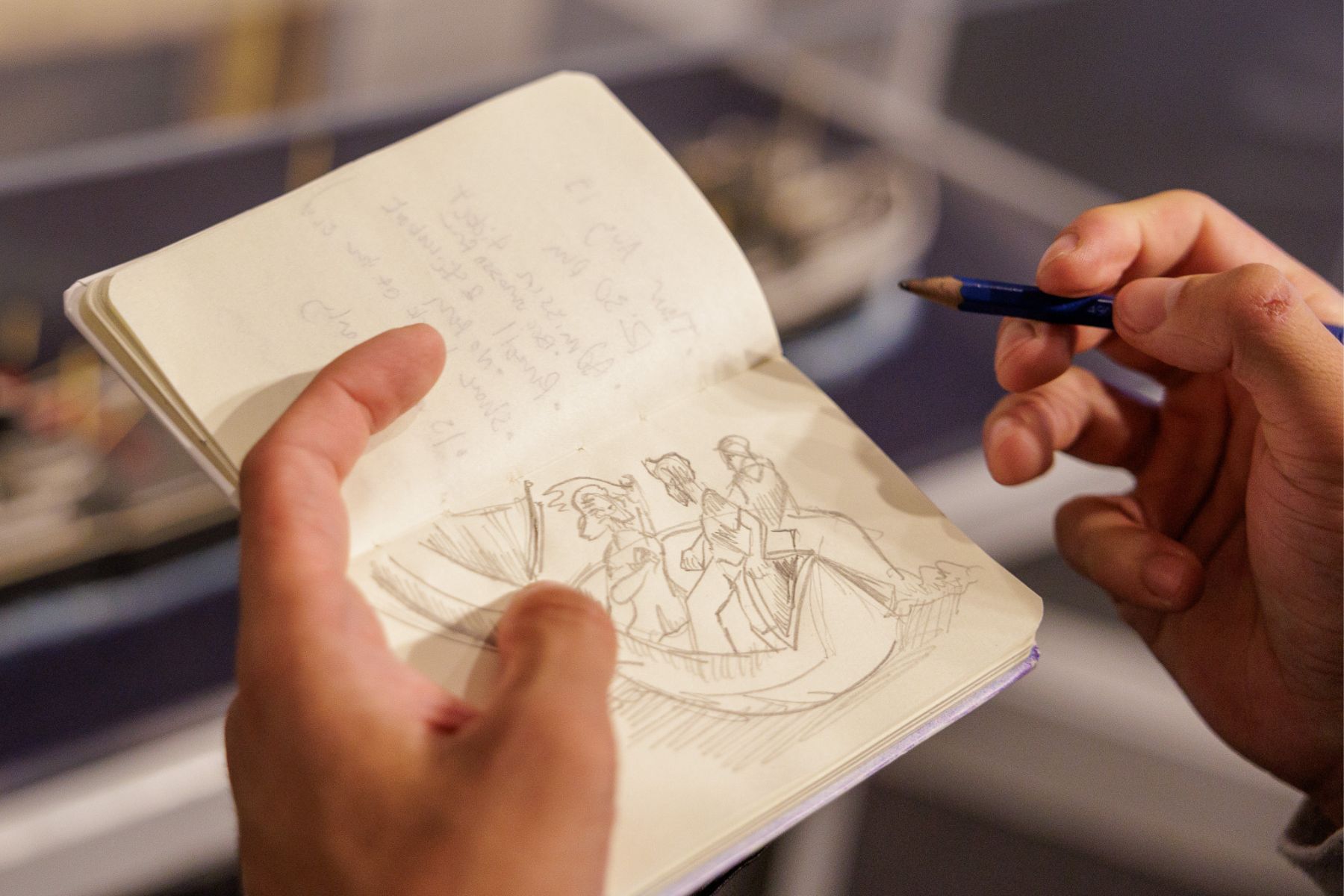 Close-up of a person's hands holding a sketchbook and pencil, featuring a detailed drawing of figures in a whaleboat. The sketch captures movement and detail, with faint notes written on the opposite page.