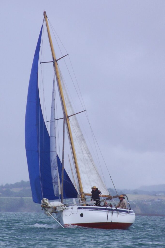 A sailboat with white and blue sails gliding through the water on a cloudy day. The boat is helmed by a small crew, and the distant shoreline is faintly visible under overcast skies.