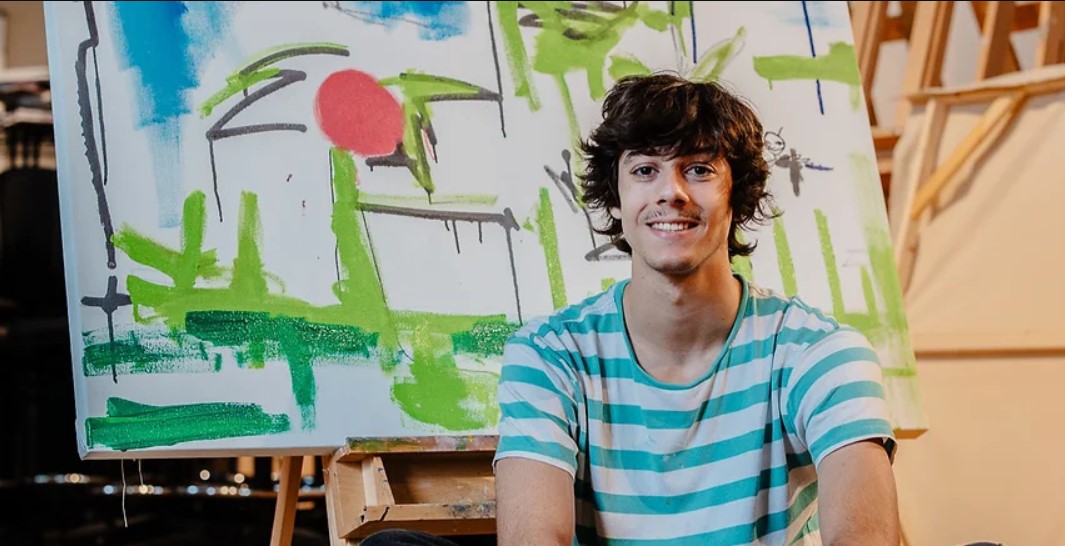 Artist Mason Terra sitting in front of an abstract painting featuring bold green, black, and red brushstrokes. He is smiling and wearing a light blue and white striped shirt, with an easel and canvas in the background of an art studio.