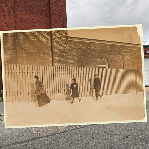 A sepia-toned historical photograph of people walking along a fence, overlaid on a modern-day photograph of the same location. The original 1912 image by Lewis W. Hine depicts Belleville Road and Nashawena Mills in New Bedford, discovered by Carl Simmons. The overlay highlights the contrast between past and present architecture.