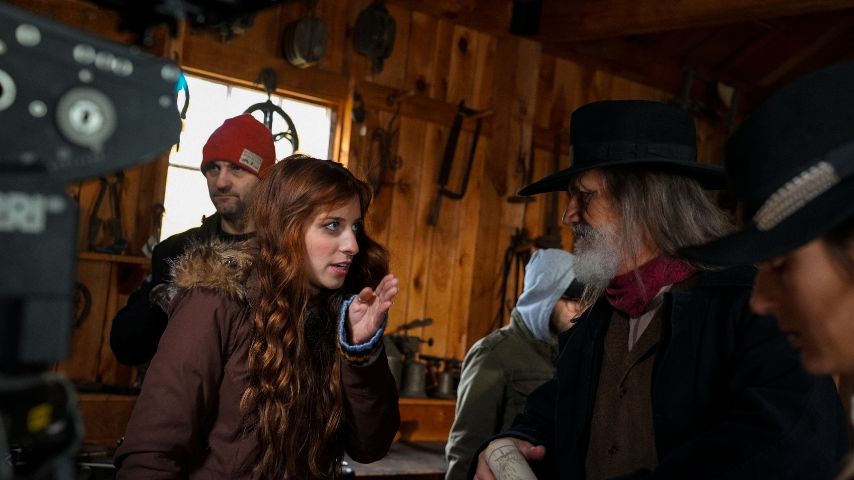 ilmmaker Alyssa Botelho, a young woman with long, wavy red hair, gestures while speaking on a film set inside a rustic wooden cabin. She wears a brown winter coat and appears engaged in conversation with actors dressed in historical costumes, including an older man with a gray beard wearing a black hat and scarf. A crew member in a red beanie stands in the background, and a professional camera is visible in the foreground.