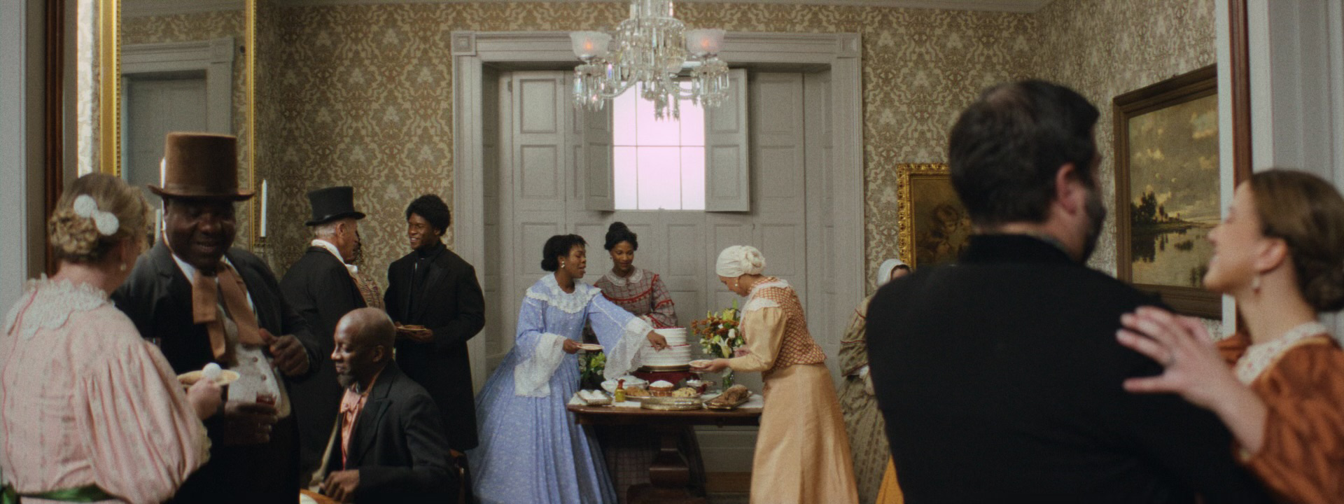 A period-style party scene with diverse guests dressed in 19th-century attire inside an elegant room. In the foreground, a man in a dark suit dances with a woman in a brown dress. To the left, a man wearing a top hat and cravat laughs while conversing with others. In the background, women in bonnets and gowns serve food from a lavish table, while additional guests engage in conversation. The room features ornate wallpaper, framed paintings, and a chandelier, evoking a historical setting.